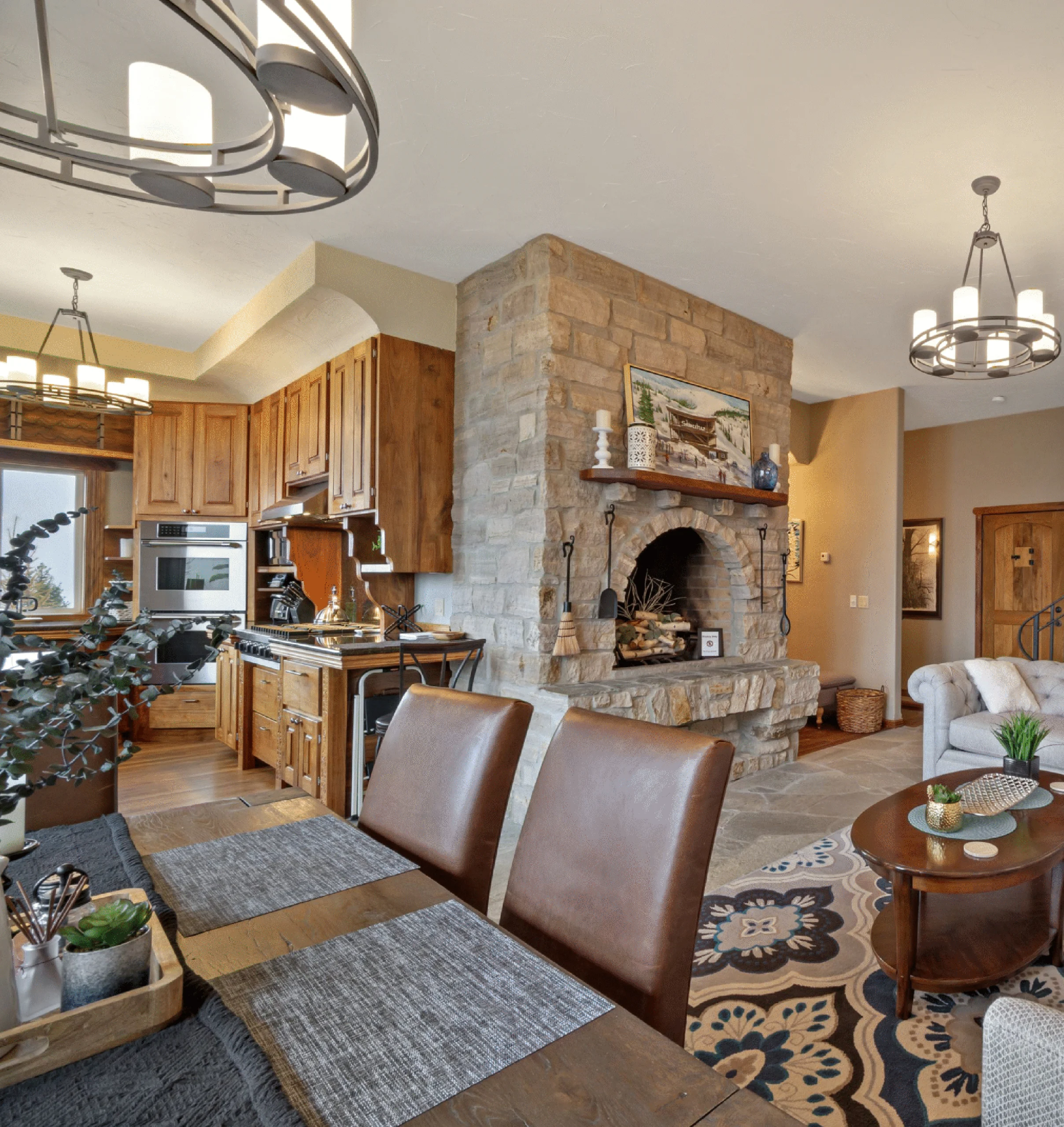 Open-plan kitchen and living area showing a stone fireplace, wood cabinets, and two circular chandeliers overhead.