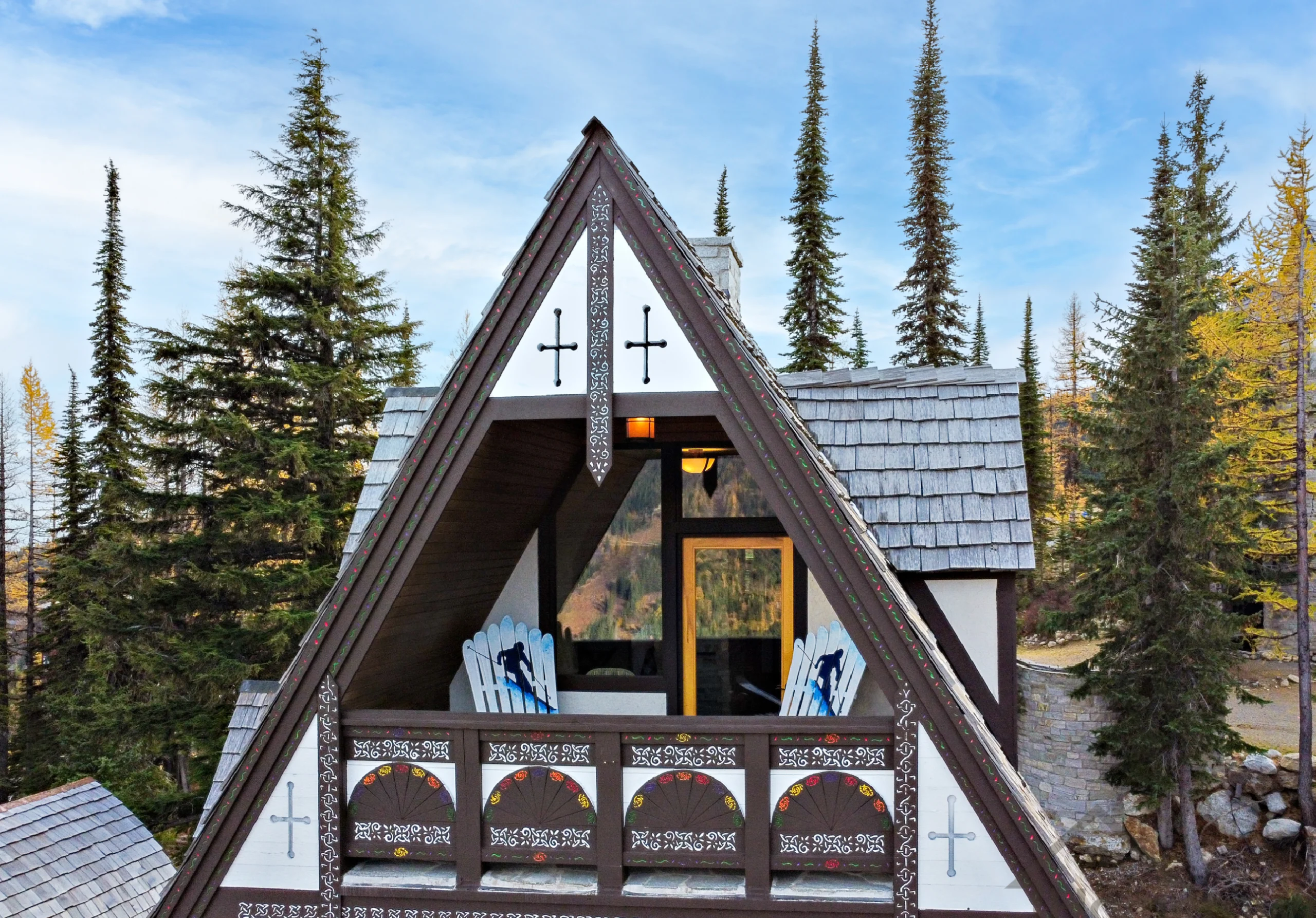 A wooden A-frame cabin with decorative white carvings along the roof edges, set among tall evergreen trees under a blue sky.