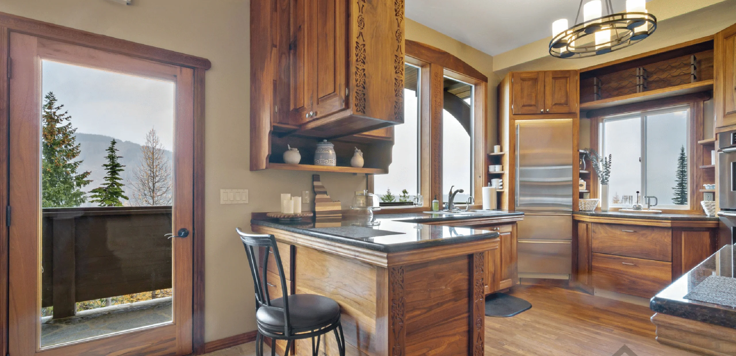 Rustic wood kitchen with carved panels, an island, and mountain trees visible through large windows/door.
